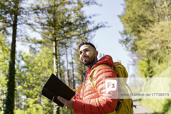 Man wearing backpack standing with diary in forest on sunny day