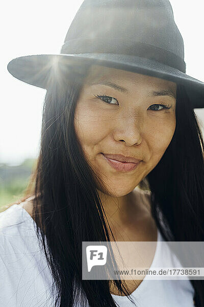 Lächelnde junge Frau mit schwarzem Haar und Hut an einem sonnigen Tag