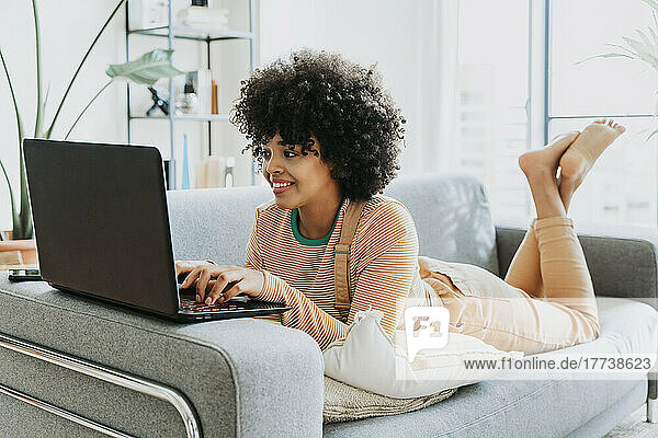 Smiling woman using laptop lying on sofa at home