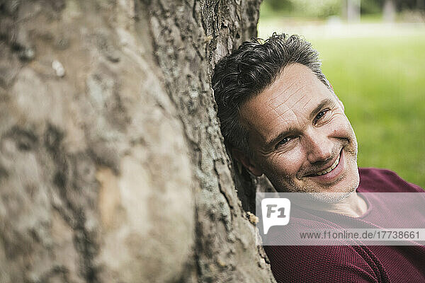 Smiling mature man leaning on tree trunk at park