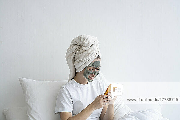 Happy woman wearing facial mask using smart phone in bedroom