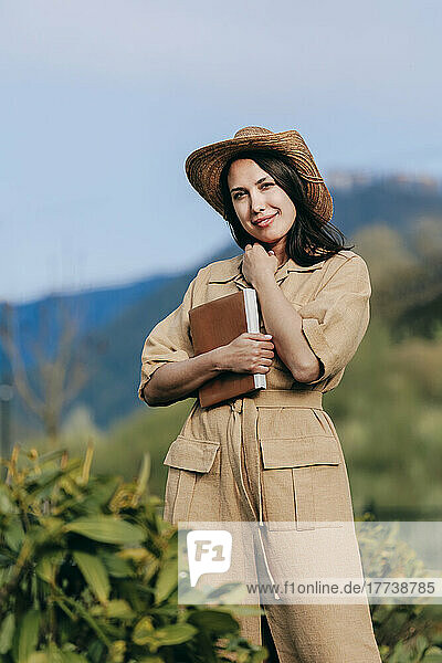 Smiling woman wearing hat standing with book in nature