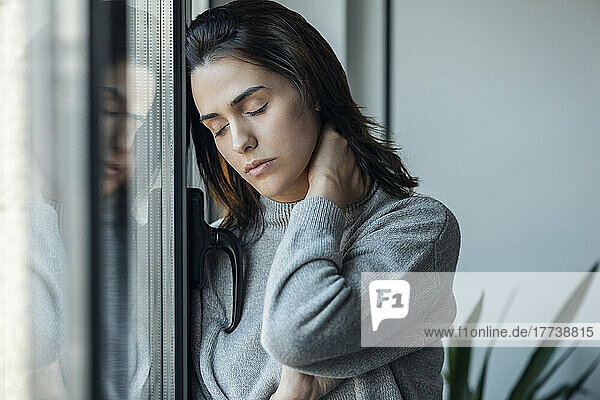 Tired woman with eyes closed standing by window at home