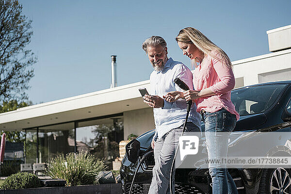 Smiling man showing mobile phone to woman holding electric plug on sunny day
