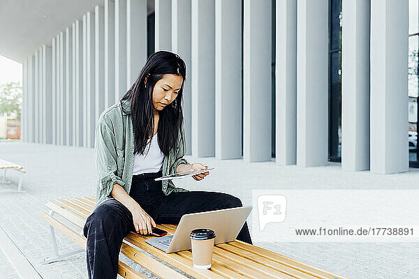 Young freelancer using tablet PC sitting with laptop and reusable coffee on bench