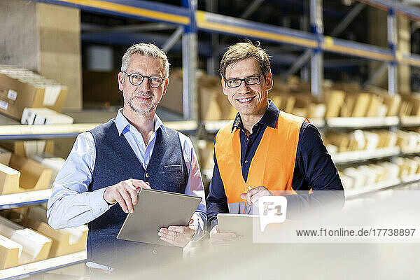 Smiling manager and worker wearing eyeglasses standing with tablet PC in warehouse