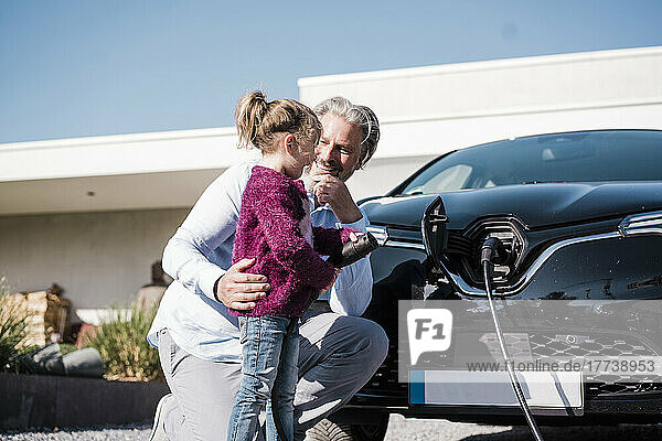 Happy girl holding electric plug standing by father in front of car on sunny day
