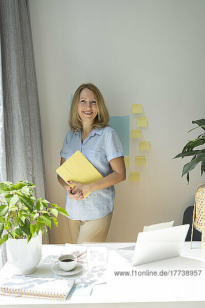 Smiling blond freelancer with tablet PC standing in home office