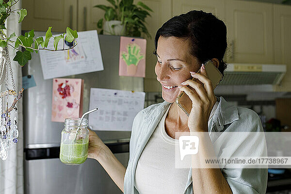 Smiling woman holding juice jar talking on mobile phone at home