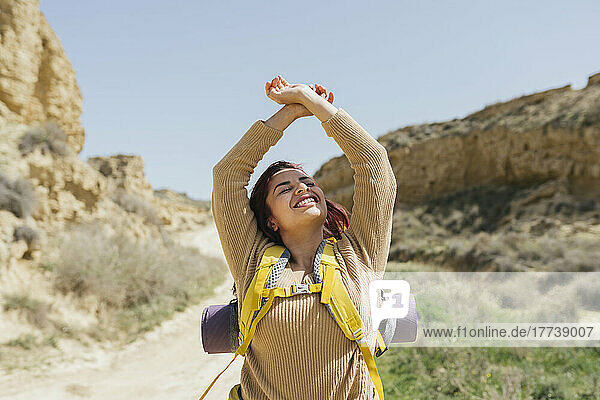 Smiling woman wearing backpack standing with arms raised on sunny day