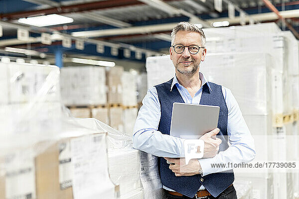 Smiling manager wearing eyeglasses standing with tablet PC by box in warehouse