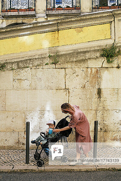 Mother feeding son sitting in baby stroller on footpath