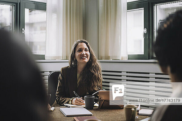 Glückliche Geschäftsfrau mit Tagebuch am Konferenztisch in einem kreativen Büro