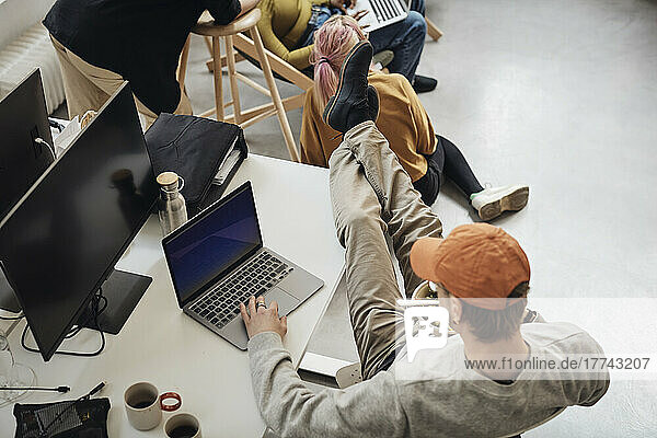 High angle view of male programmer with feet up on desk using laptop while eating lunch in office