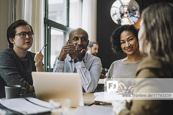 Lächelnde männliche und weibliche Kollegen betrachten eine Geschäftsfrau  die am Schreibtisch in einem kreativen Büro einen Plan bespricht