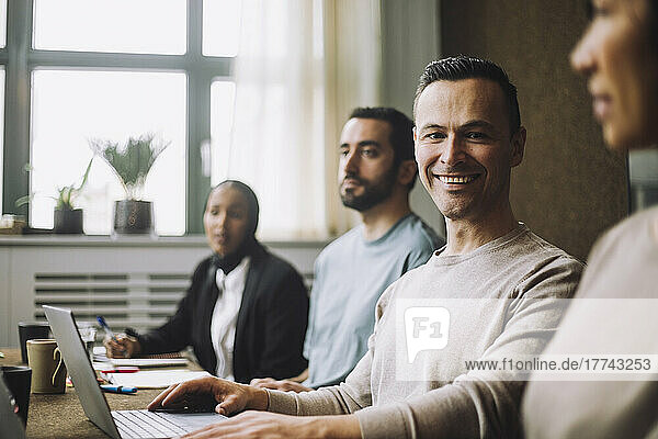 Happy mature businessman with laptop sitting by colleagues in meeting room