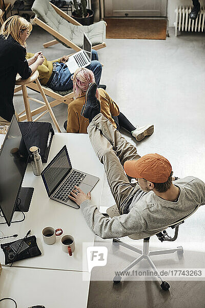 Männlicher Unternehmer mit hochgelegten Füßen auf dem Schreibtisch  der einen Laptop benutzt  während seine Kollegen im Büro eines Technologie-Start-ups diskutieren