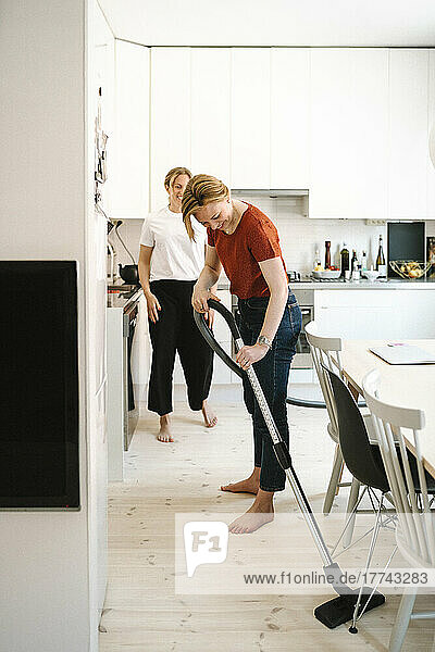 Frau reinigt Boden mit Staubsauger  während Freundin in der Küche steht