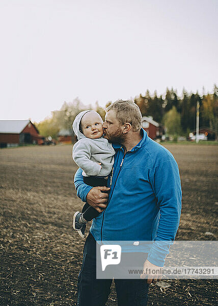Vater küsst Kleinkind Sohn stehen auf dem Bauernhof bei Sonnenuntergang