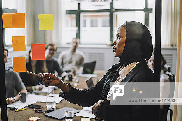 Young businessman wearing headscarf explaining adhesive notes to colleagues during meeting in creative office