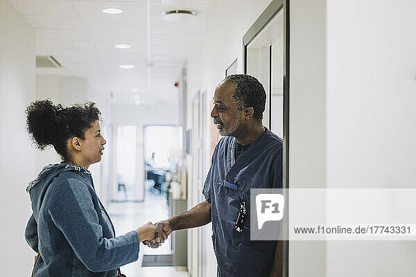 Smiling doctor greeting female patient with handshake while standing at doorway in hospital
