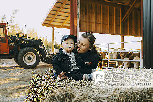 Mutter betrachtet ihren Sohn  der auf einem Heuballen auf einem Bauernhof sitzt