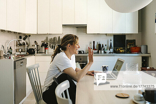 Happy female freelancer waving on video call through laptop at table in kitchen