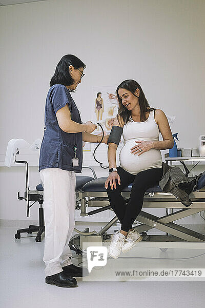 Ärztin in voller Länge  die den Blutdruck einer schwangeren Frau auf einer Trage sitzend in einer medizinischen Klinik überprüft