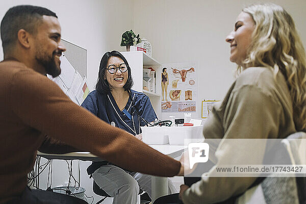 Glückliche Gynäkologin betrachtet einen lächelnden Mann  der den Bauch einer schwangeren Frau in der Klinik berührt