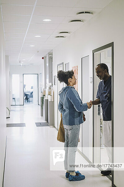 Full length of smiling doctor greeting female patient standing in corridor at hospital