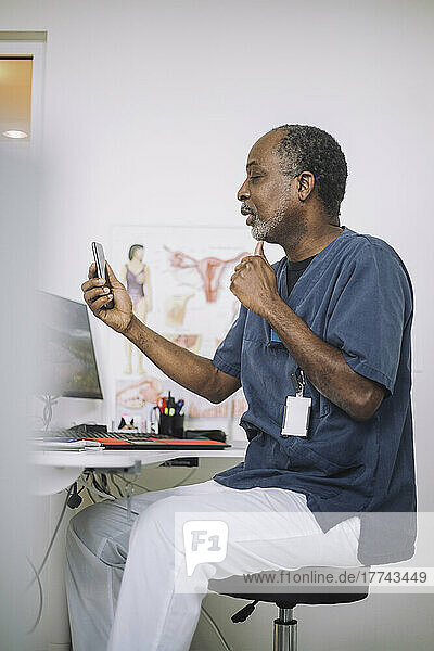Männlicher Arzt bei der Online-Konsultation über das Mobiltelefon  während er am Schreibtisch in der Klinik sitzt