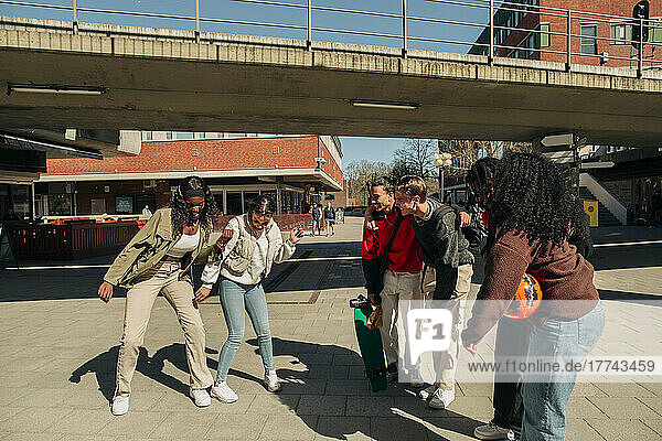 Multiracial friends watching at young women dancing together on street during sunny day