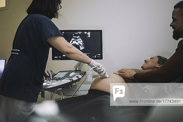 Lächelndes Paar  das auf den Bildschirm schaut  während ein Gynäkologe im Krankenhaus Ultraschalluntersuchungen durchführt