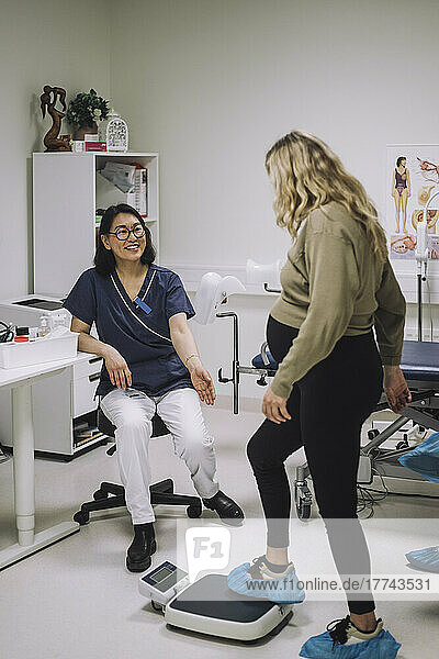 Lächelnde Ärztin in voller Länge  die eine schwangere Frau auf der Waage in einer medizinischen Klinik betrachtet