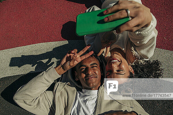 Friends making peace sign while taking selfie through smart phone lying in playground
