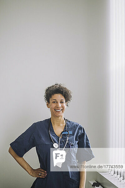Portrait of happy female doctor standing with hand on hip against white wall in hospital