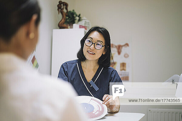 Eine lächelnde Mitarbeiterin des Gesundheitswesens zeigt eine Tabelle zur In-vitro-Fertilisation  während sie mit einem Patienten in der Klinik spricht.