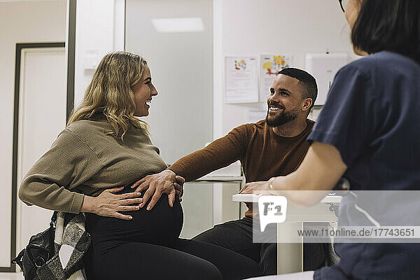 Glücklicher Mann  der den Bauch einer schwangeren Frau berührt  während er mit einer Ärztin in einer medizinischen Klinik sitzt