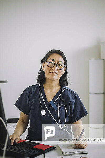 Portrait of smiling female doctor wearing eyeglasses writing while sitting at desk in medical clinic