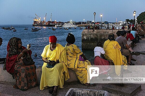 Frauen in bunten Tüchern sitzen an der Promenade des Fodharani Park  hinten Hafen  Dämmerung  Stone Town  Altstadt  Unguja  Sansibar  Tansania  Afrika