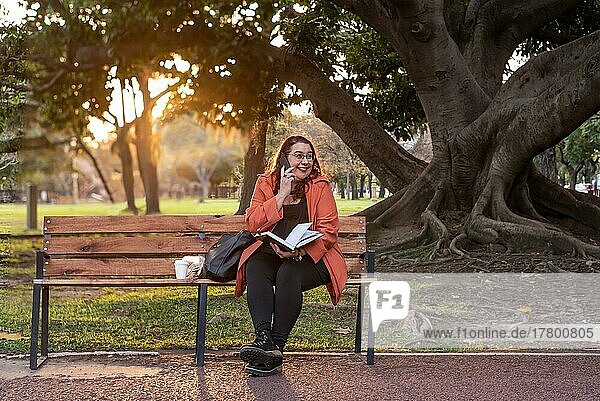 Eine leitende Frau in Übergröße arbeitet im Freien. Sie hält ein Notebook und führt ein Telefongespräch  während sie lacht und auf einer Bank in einem öffentlichen Park sitzt
