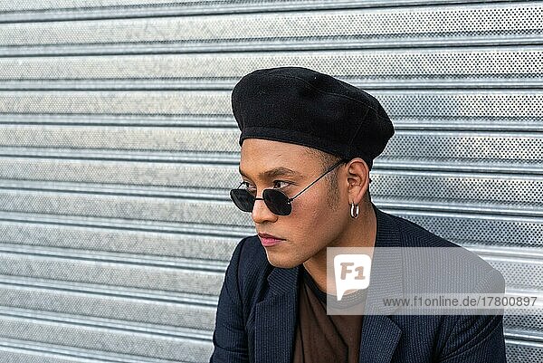 Young latin gay man with make-up on wearing a fashionable hat and looking over sunglasses  before a metal curtan. LGBT