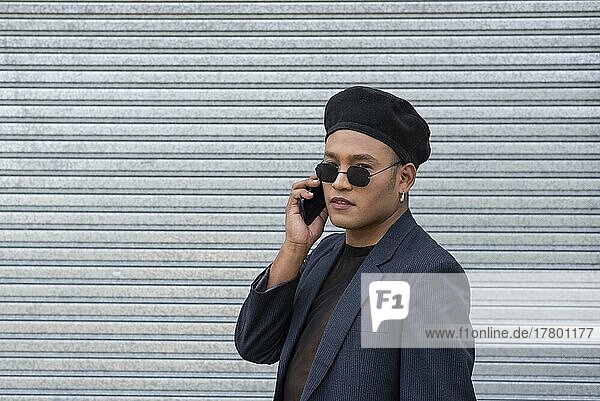 Young Latin gay man wearing a fashionable hat and sunglasses  talking on his smartphone  before a metal curtain background. LGTB. Copy space