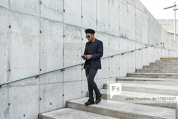 Young Latin gay man wearing a fashionable hat and sunglasses  walking down the stairway while texting on his smartphone  before a white background. LGTB. Copy space