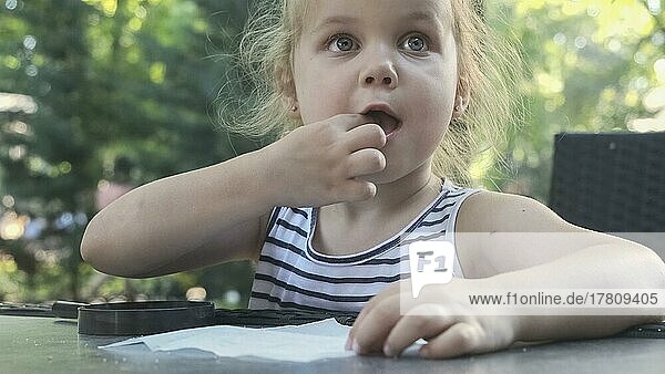 Kleines Mädchen schmeckt Salz. Close-up-Porträt von blonden Mädchen nimmt Salz aus Serviette mit dem Finger und schmeckt es  während in Straßencafé im Park sitzen. Odessa  Ukraine  Europa