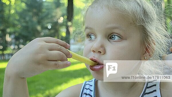 Kleines Mädchen isst Pommes frites. Nahaufnahme von blonden Mädchen nimmt Kartoffelchips mit ihren Händen und probiert sie sitzen in Straßencafé auf dem Park. Odessa Ukraine