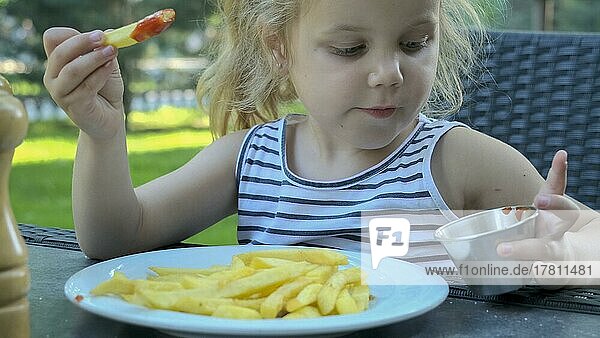 Kleines Mädchen isst Pommes frites. Nahaufnahme von blonden Mädchen nimmt Kartoffelchips mit ihren Händen und probiert sie sitzen in Straßencafé auf dem Park. Odessa Ukraine