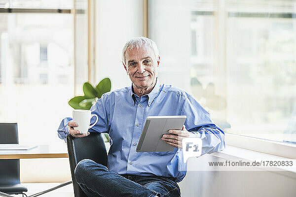 Smiling businessman with coffee cup and tablet PC at work place