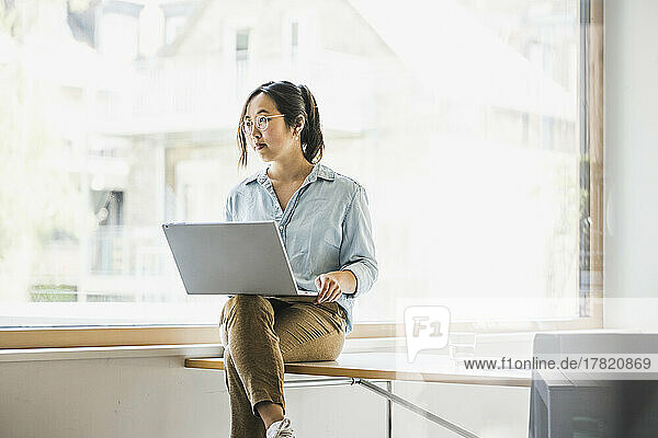 Businesswoman with laptop sitting on desk by window