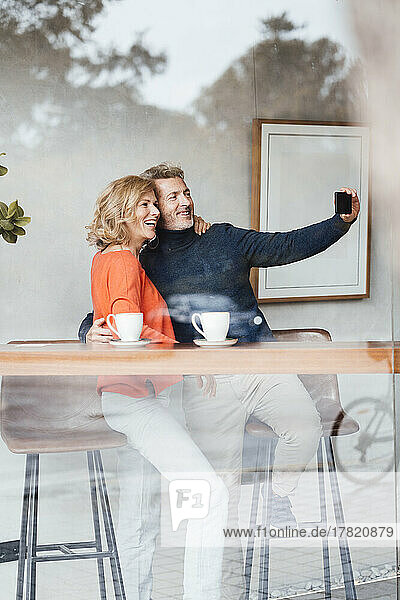 Smiling man with woman taking selfie in cafe seen through glass window